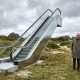 Rolltreppe mitten in den Dünen von Kampen, daneben ein älteres Paar und ein Bauarbeiter mit Bauplan, im Hintergrund Reetdachhäuser